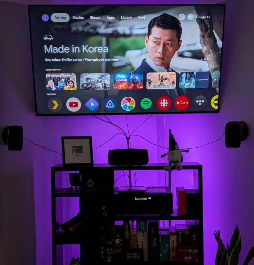 Wall-mounted Sony TV displaying the Google TV home screen with purple LED bias lighting, flanked by bookshelf speakers and a home theater receiver on a shelf below.